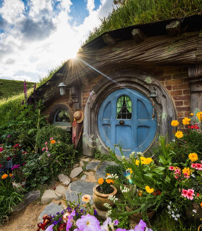 Blue round door of a hobbit house surrounded by colorful flowers and green grass with sun shining in the background.