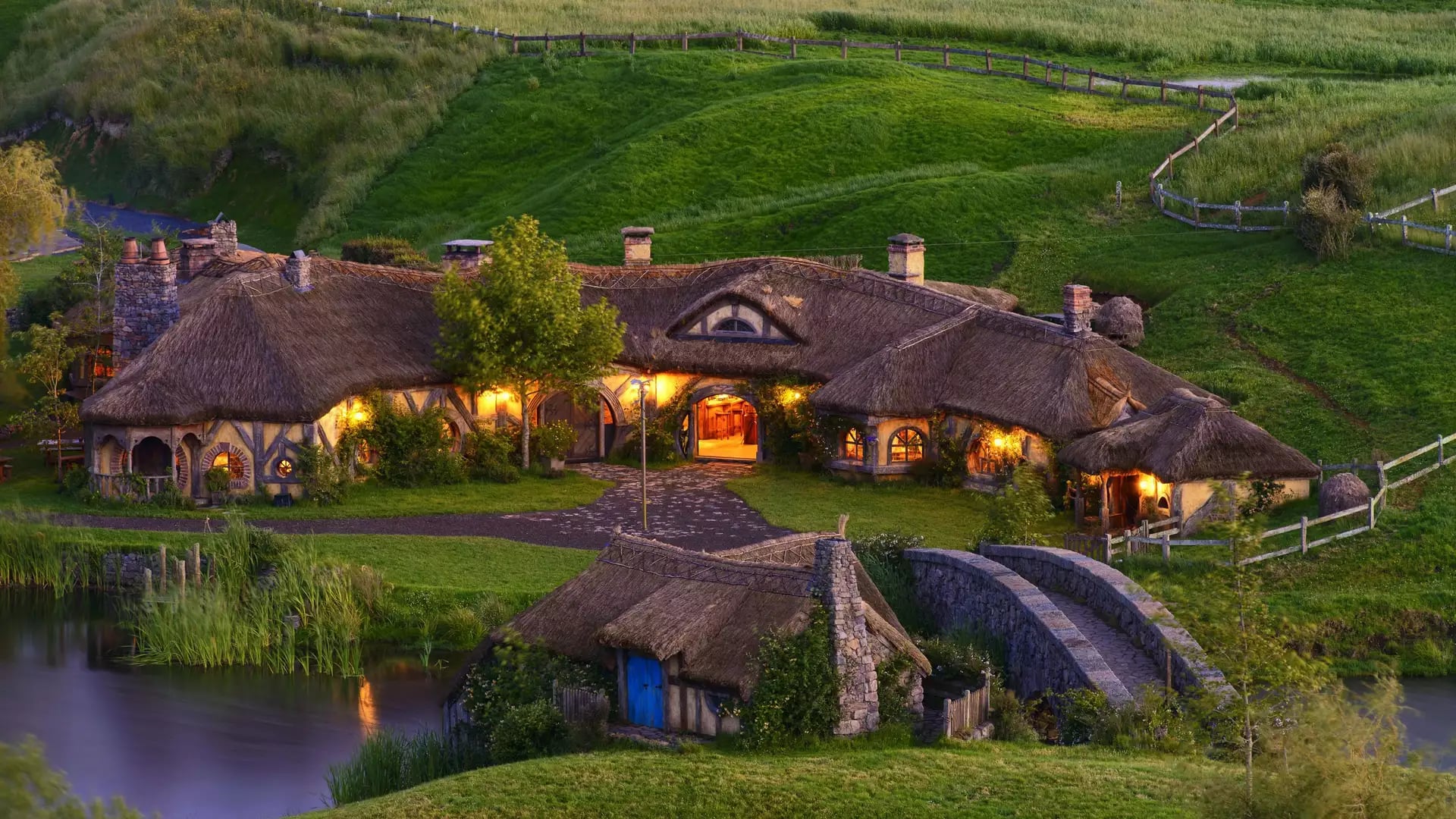 Thatched roof hobbiton cottage and stone bridge surrounded by green hills at sunset near a pond