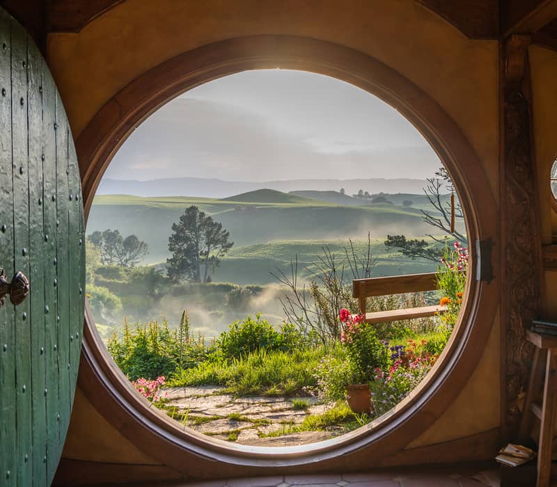 Inside view of a hobbit hole with a round door open to a green garden and rolling hills beyond.