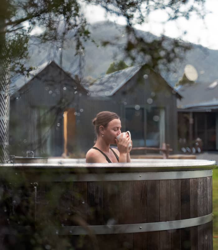 Woman in an outdoor wooden hot tub sipping from a cup with cabins and mountains behind her.