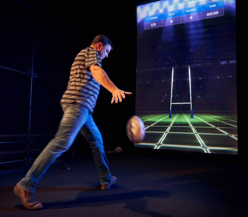 Man kicking rugby ball towards a digital rugby goal on a large indoor projection screen.