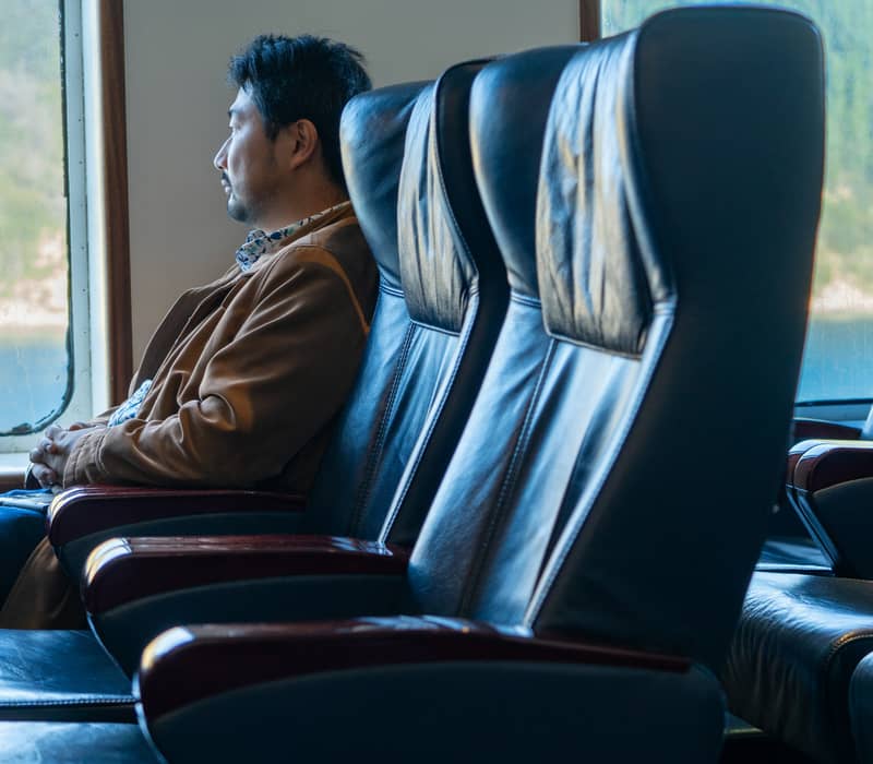 Man sitting in a boat lounge looking out a window at forest and water scenery.