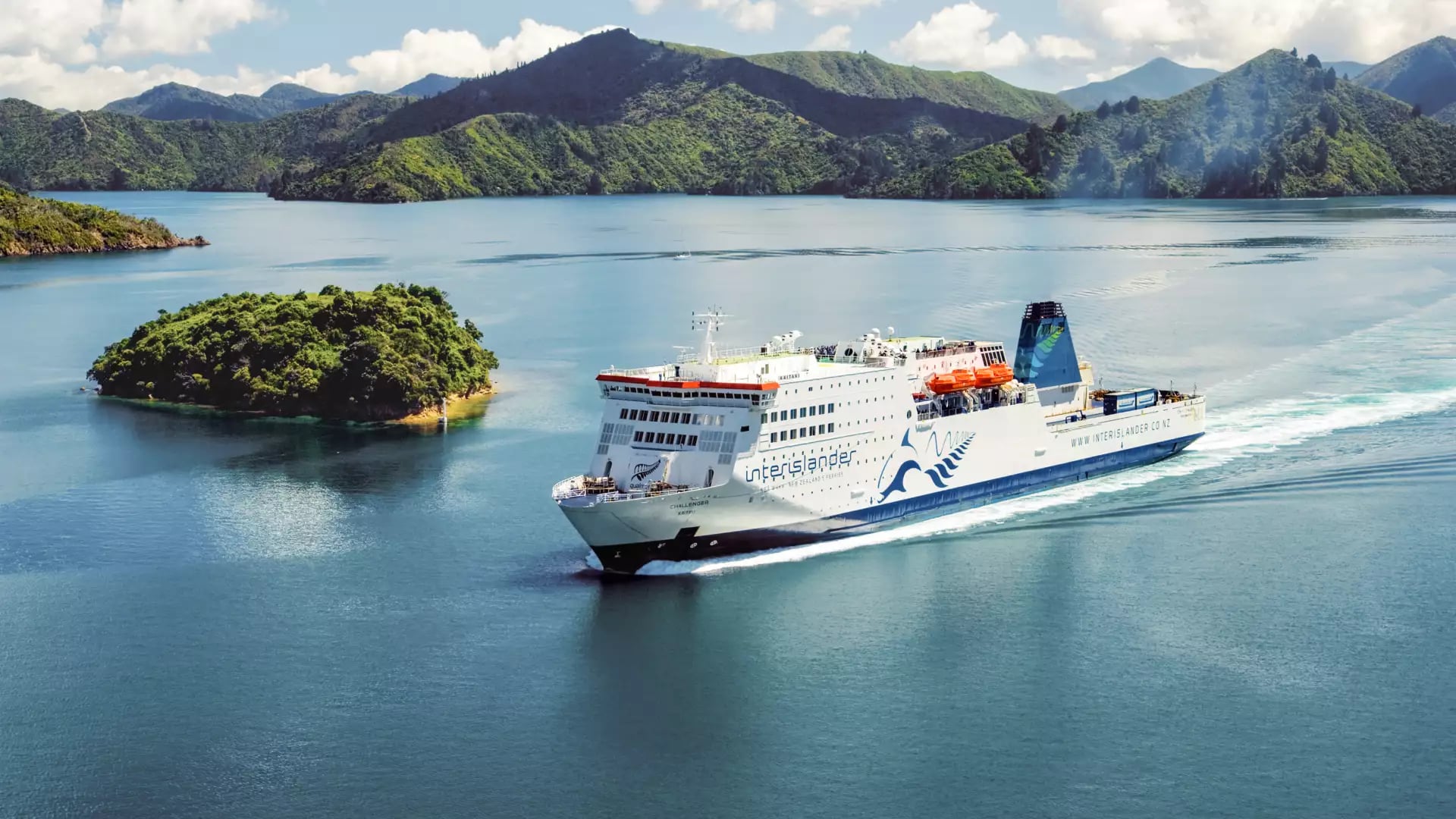 A large ferry sails through blue waters with green islands and mountains in the background under a partly cloudy sky.