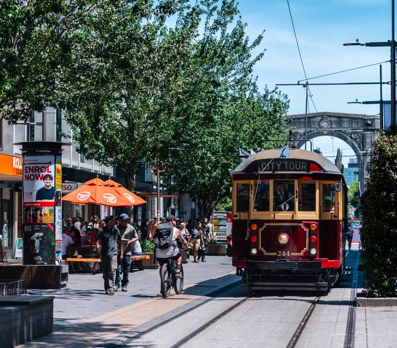 Vintage city tour tram on a sunny street with pedestrians, trees, and cafes.
