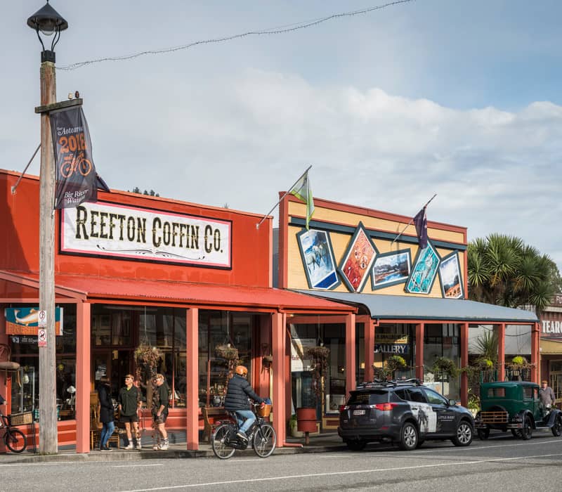 People and bicycles outside shops on Reefton Street with cars parked along the road during the day.