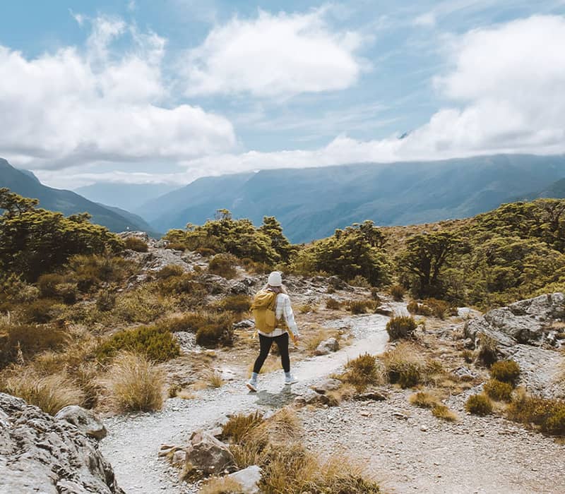 Person hiking on rocky mountain trail with trees and mountains in the background under cloudy sky.