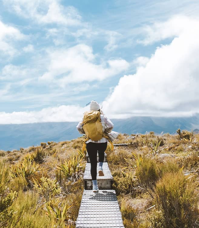 Person hiking on a boardwalk trail in the mountains with a yellow backpack and cloudy sky.