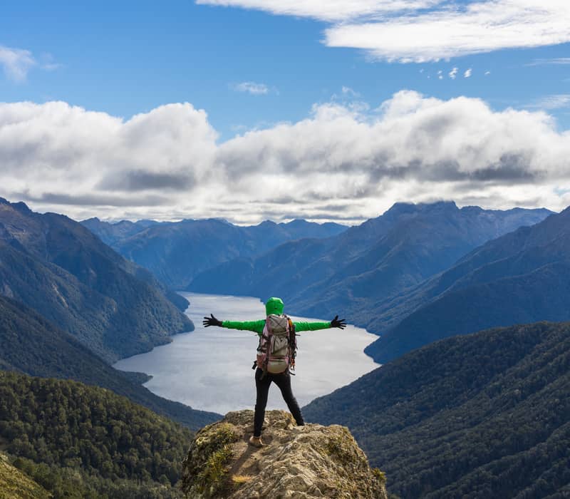 Hiker in green jacket stands on rock with arms outstretched overlooking lake and mountain range under cloudy sky.