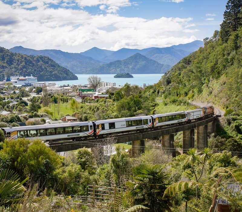 Passenger train crosses a viaduct with green hills and mountains in the background near water