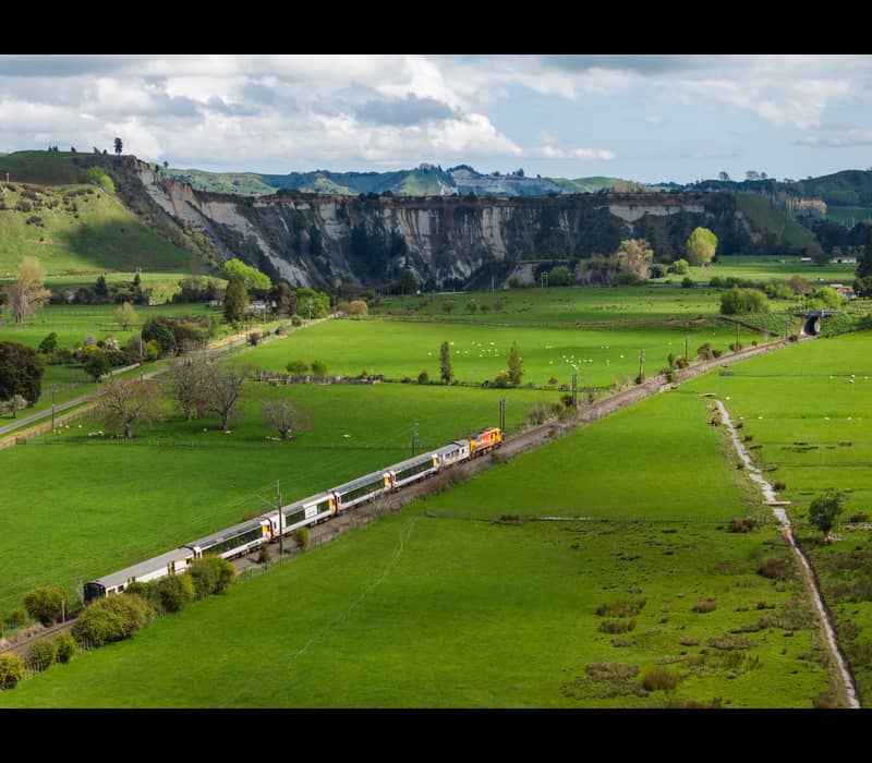 Northern Explorer train traveling on railway track through green farmland near Rangitikei Viaduct with hills behind.