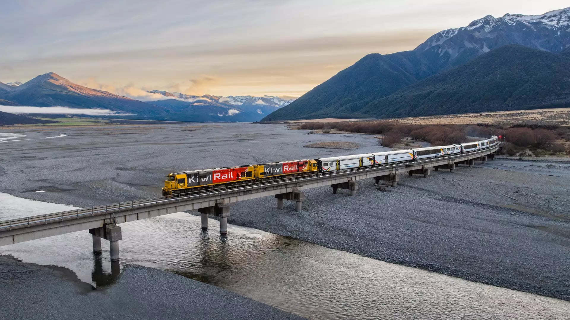Aerial view of a KiwiRail train on a bridge over river with mountains and clouds at sunset.