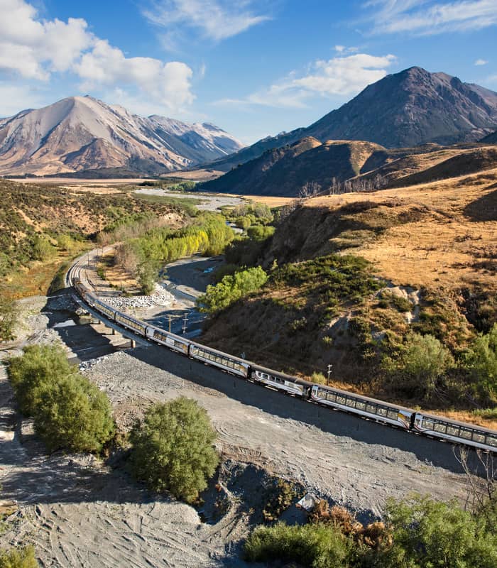 TranzAlpine train crossing a river bridge in a mountainous valley under blue sky and clouds