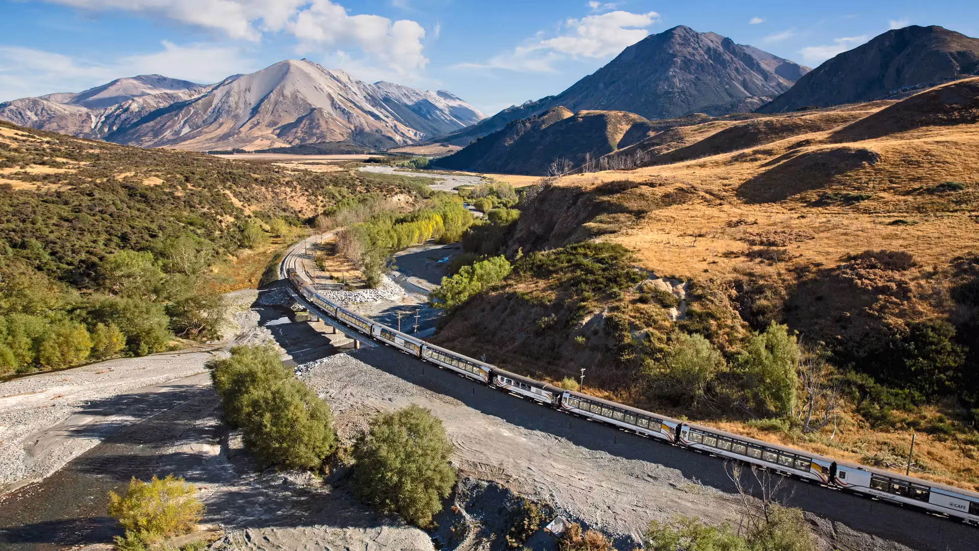 TranzAlpine train crossing a river bridge in a mountainous valley under blue sky and clouds