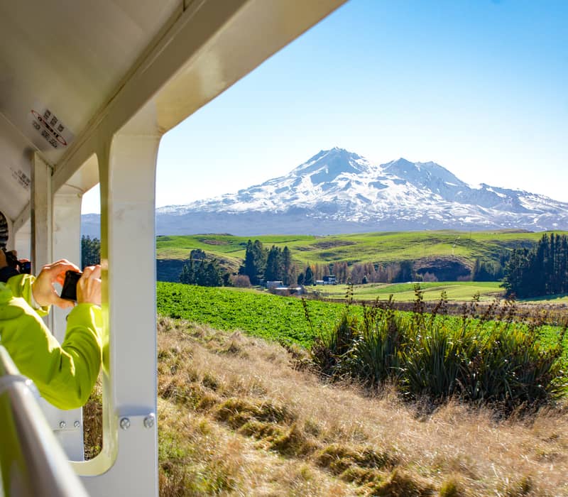 Person on train taking photo of snow-capped mountain and green fields under a clear blue sky.