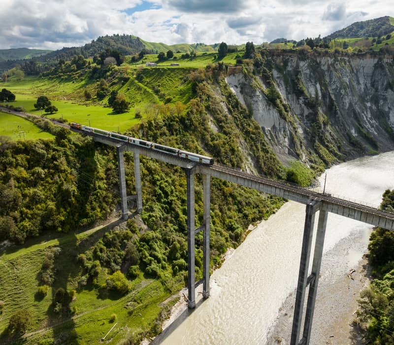 Train traveling across a high viaduct over a river surrounded by green hills and forested cliffs.