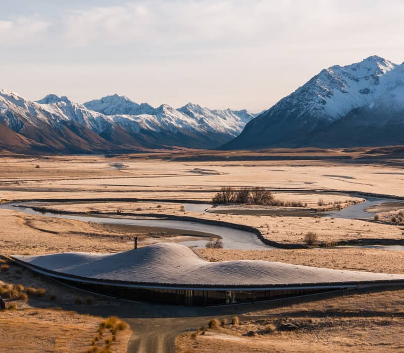Modern building in valley with winding river and snow-covered mountains under cloudy sky.