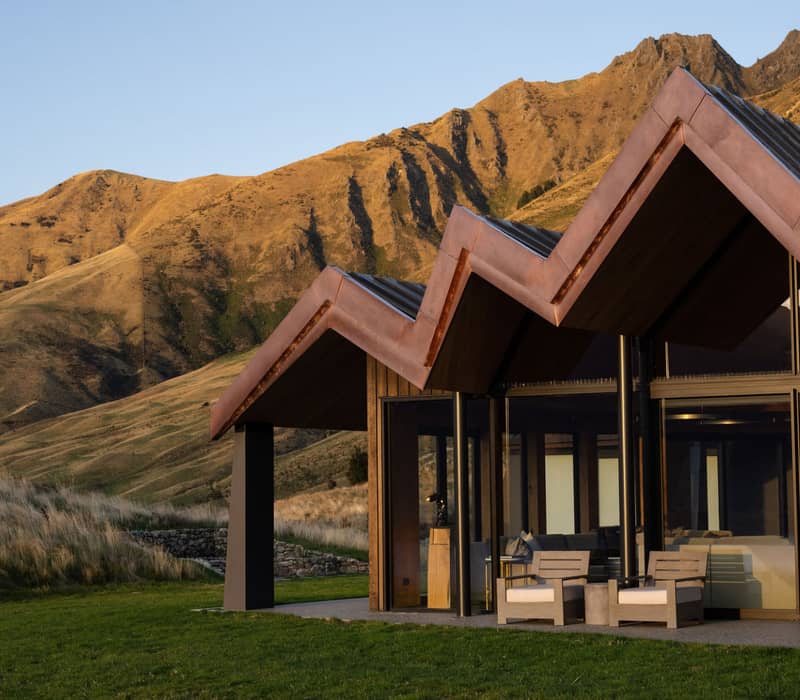 Modern house with zigzag roof and glass walls beside grassy lawn and mountains at sunset.