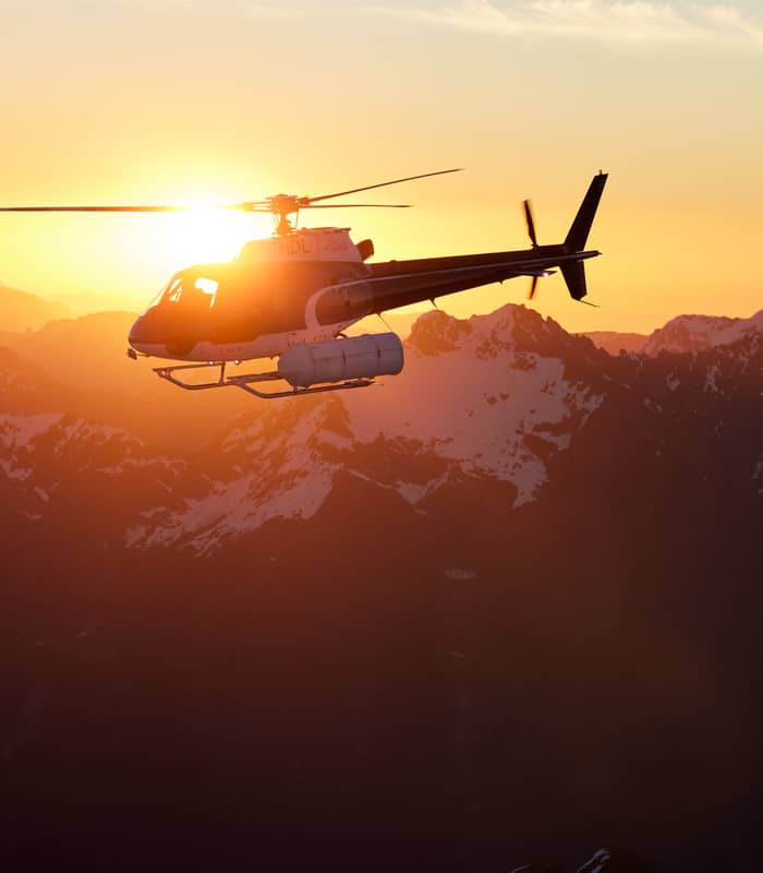 Helicopter flying above snowy mountains at sunset with bright orange sun in the background.
