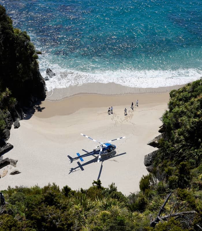 Helicopter on a sandy beach with people walking and ocean waves nearby, surrounded by green cliffs.