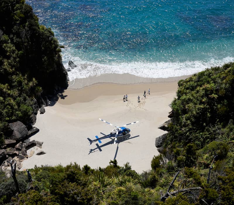 Helicopter on a sandy beach with people walking and ocean waves nearby, surrounded by green cliffs.