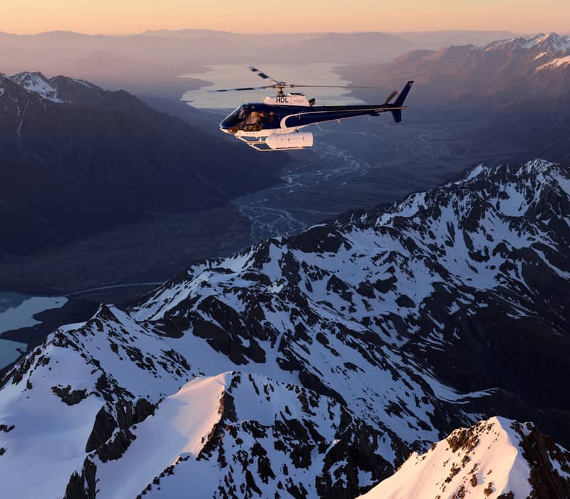 Helicopter flying over snowy mountains and glaciers at sunset with river and lake below.