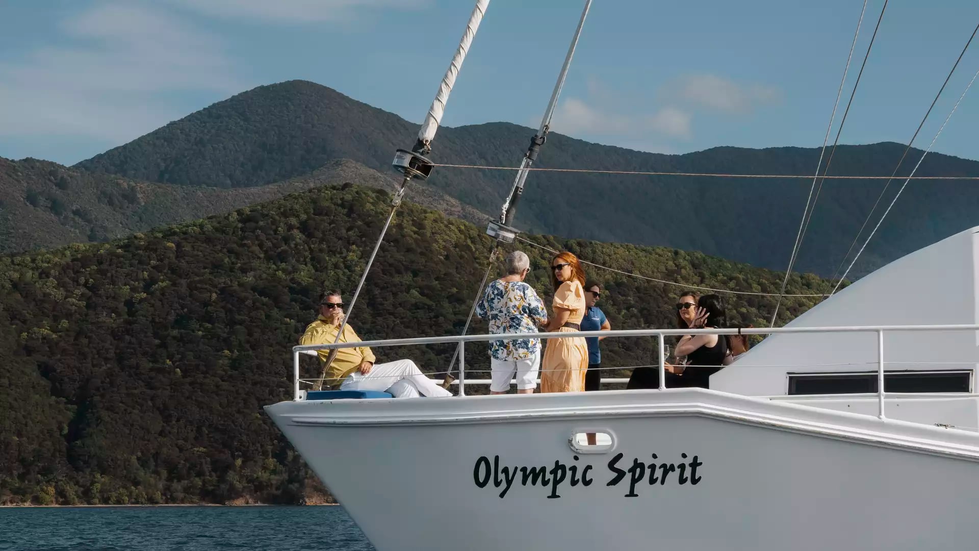 People on a sailboat named Olympic Spirit with mountains and water in the background on a sunny day.