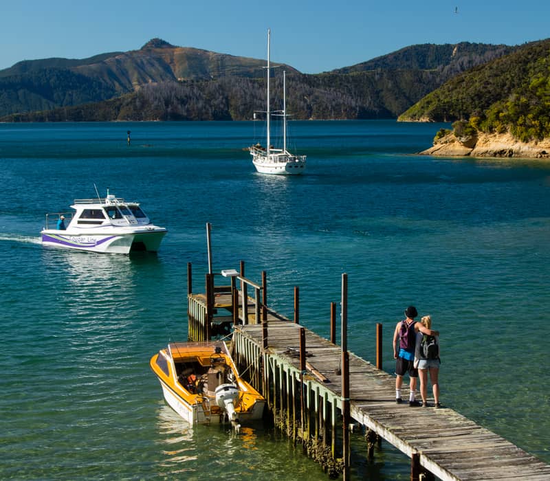 Small boat approaching dock with couple, mountains and clear water in background.