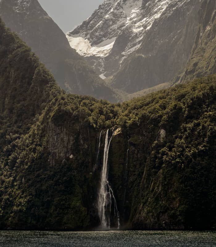 Waterfall flows down forested cliff with snowy mountain peaks behind and water in the foreground.
