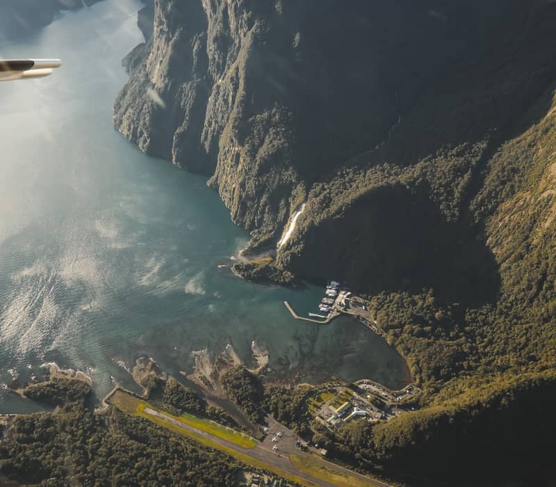 Aerial shot of Milford Sound showing mountains, runway, waterfalls, harbor, and river.