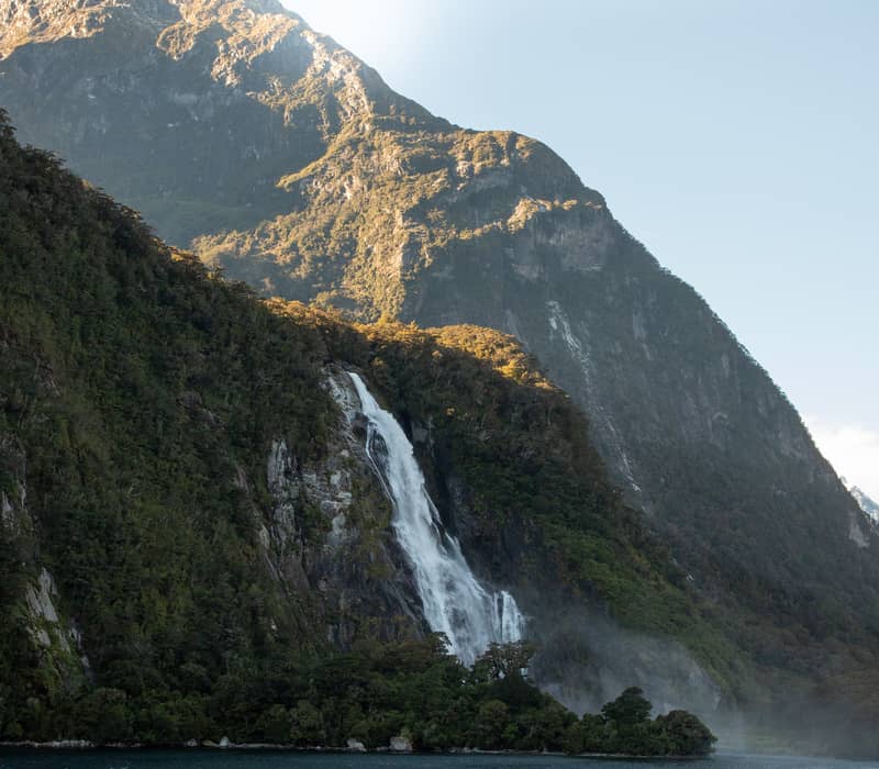 Waterfall flowing down a mountain into a river with forest and clear sky.