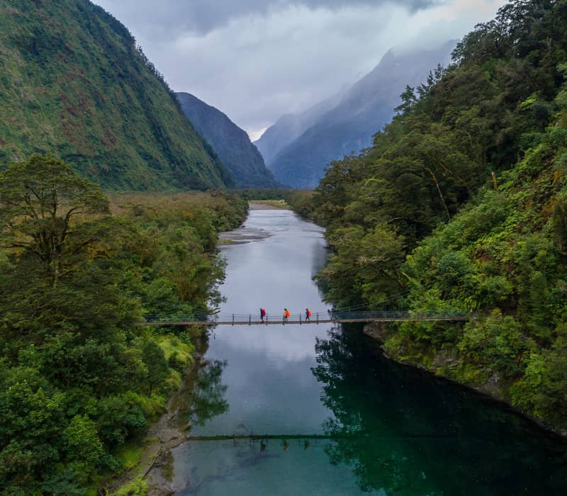 Three hikers crossing a suspension bridge over a river in a lush forested mountain valley.