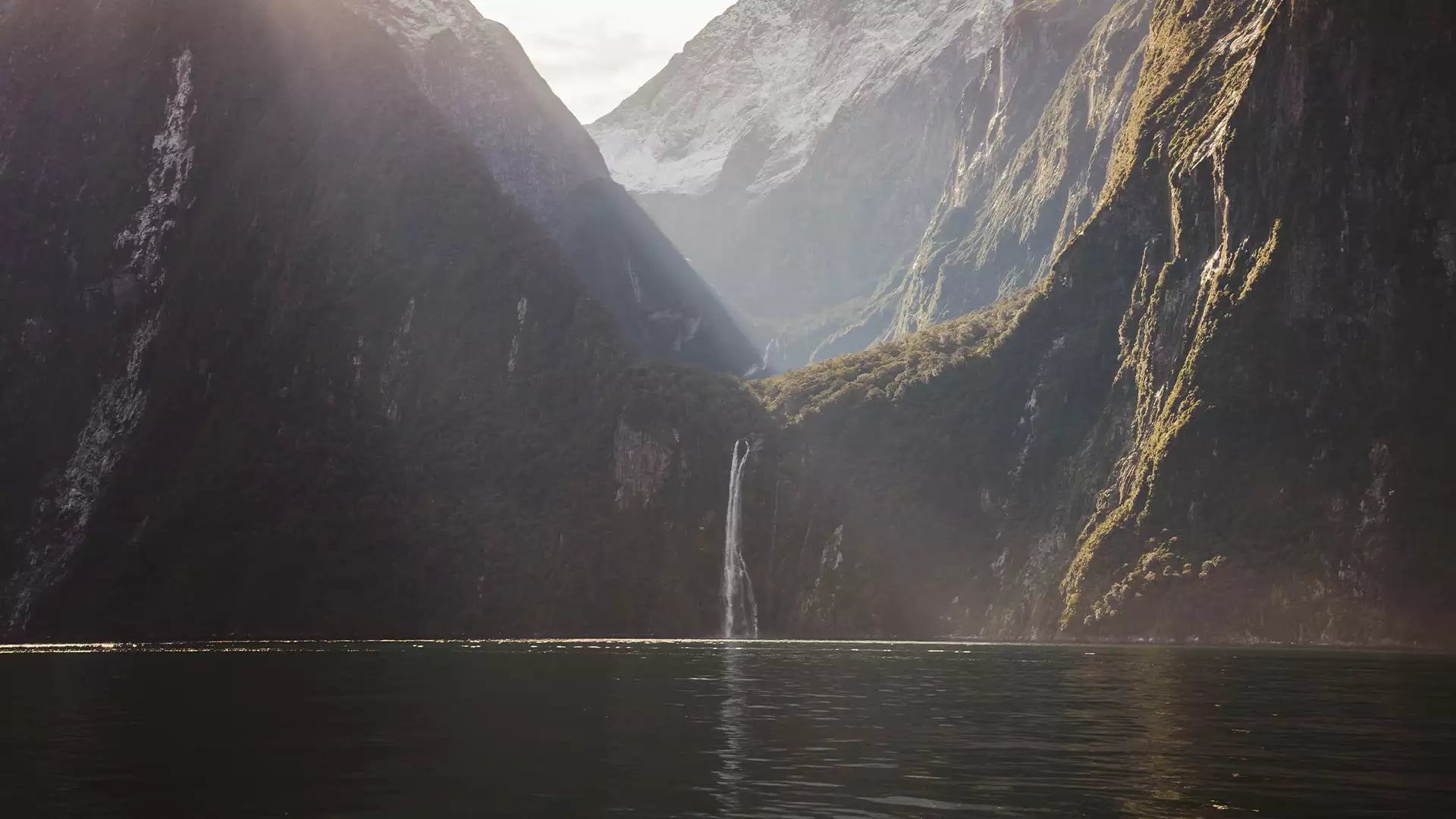 Waterfall flowing down a mountain cliff into a lake with snow-capped peaks and forest nearby.