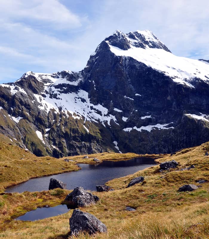 Snow-capped mountain behind a small alpine lake with grassy and rocky terrain.