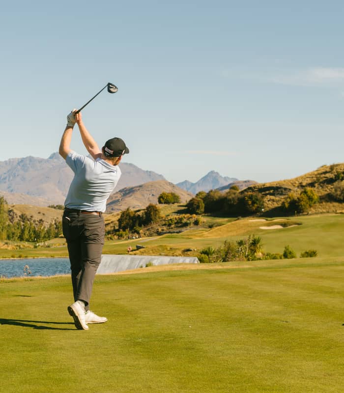 Man swinging golf club on a green course with mountains and water in the background under clear sky.