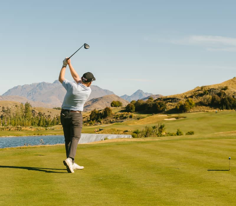 Man swinging golf club on a green course with mountains and water in the background under clear sky.