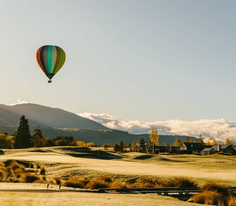 Colorful hot air balloon flying over a golf course with mountains and houses in the distance.