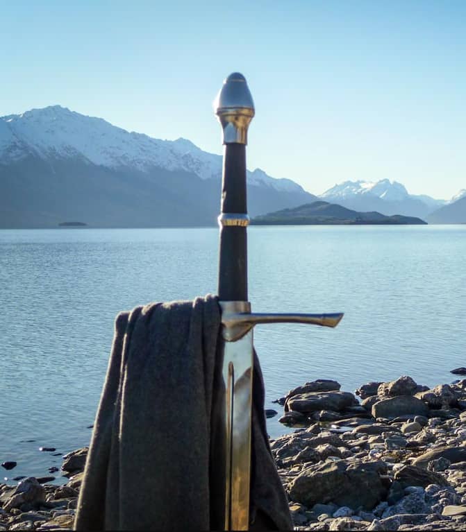 Sword with scarf on rocky shore by lake with snow-capped mountains.