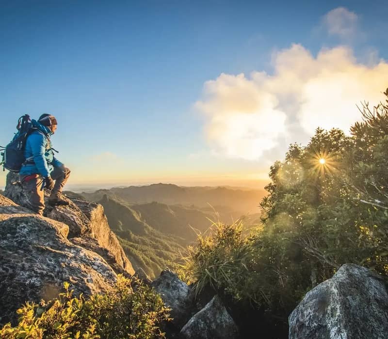 Hiker standing on rocky cliff at sunrise with mountain and forest views in distance