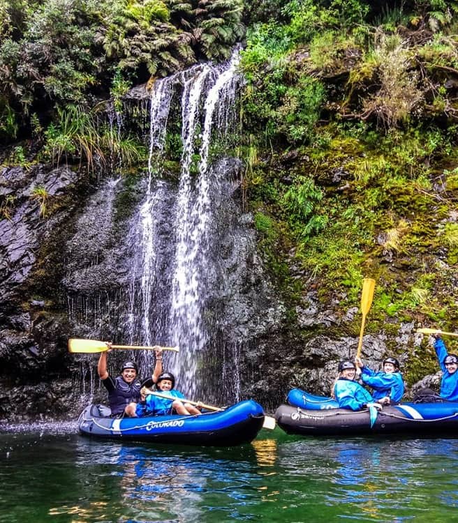 People kayaking in blue rafts near a waterfall with green forest and rocks.