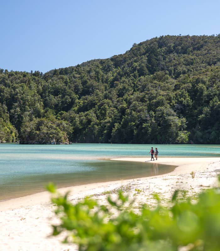 Two people walking on a sandy beach with turquoise water and green forest hills in the background under clear sky.