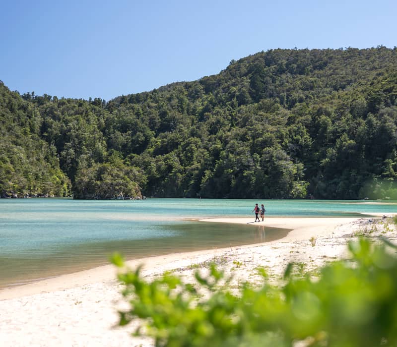 Two people walking on a sandy beach with turquoise water and green forest hills in the background under clear sky.