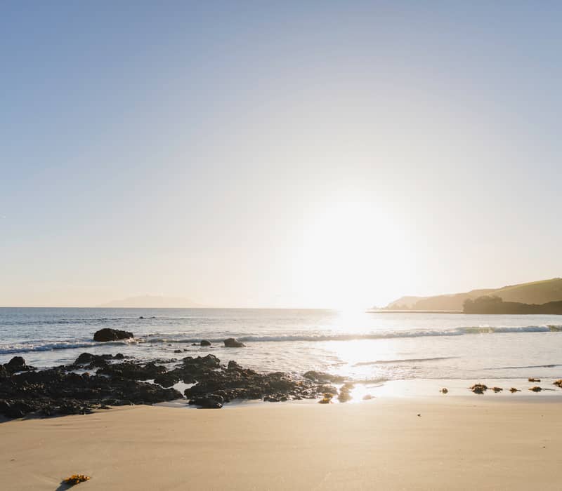 Sun setting over a sandy beach with calm waves and rocks scattered near the shore.