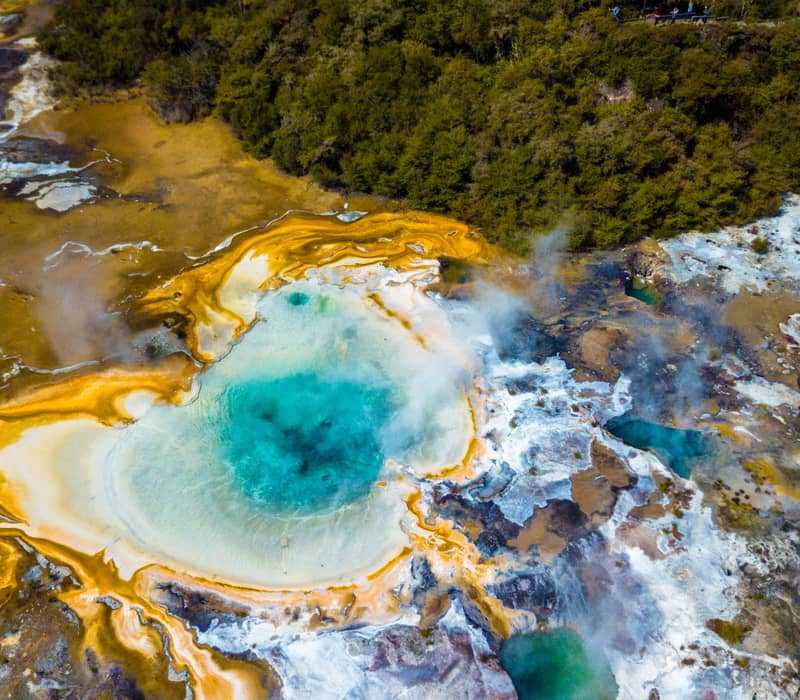 Aerial view of a colorful geothermal hot spring with orange and blue mineral deposits emitting steam near forest.