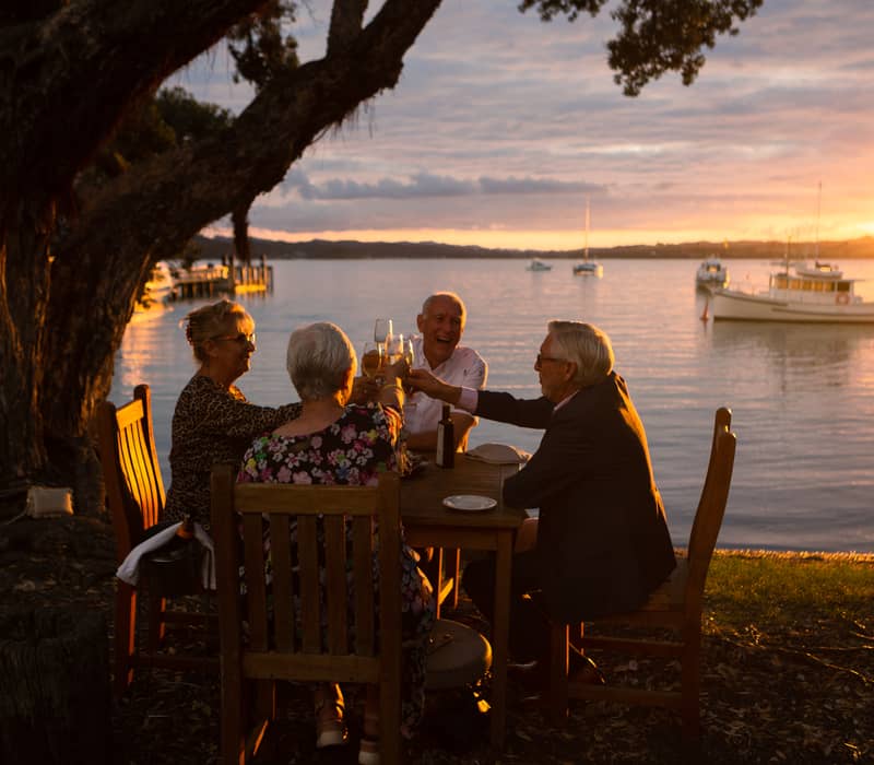 Four elderly people toasting drinks at a lakeside dinner table at sunset with boats on water.