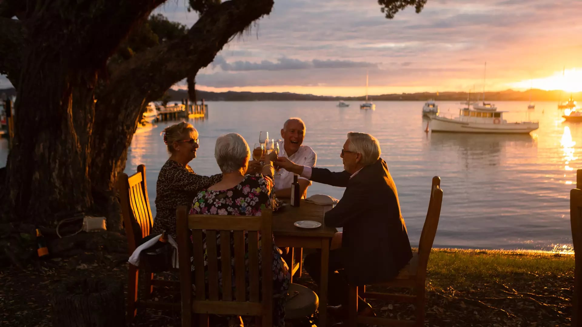 Four elderly people toasting drinks at a lakeside dinner table at sunset with boats on water.