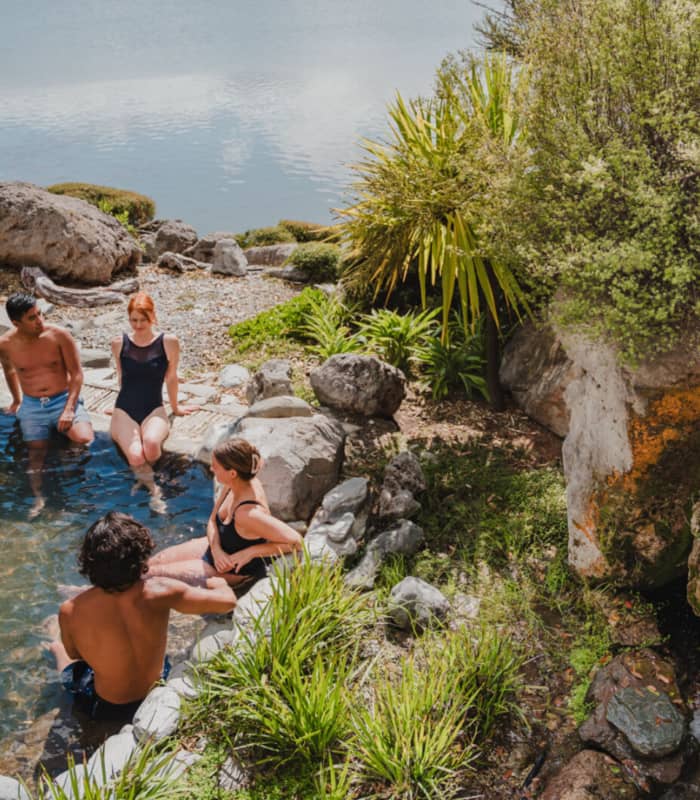Four people relaxing in an outdoor hot pool with rocks, plants, and a waterfall near a lake.