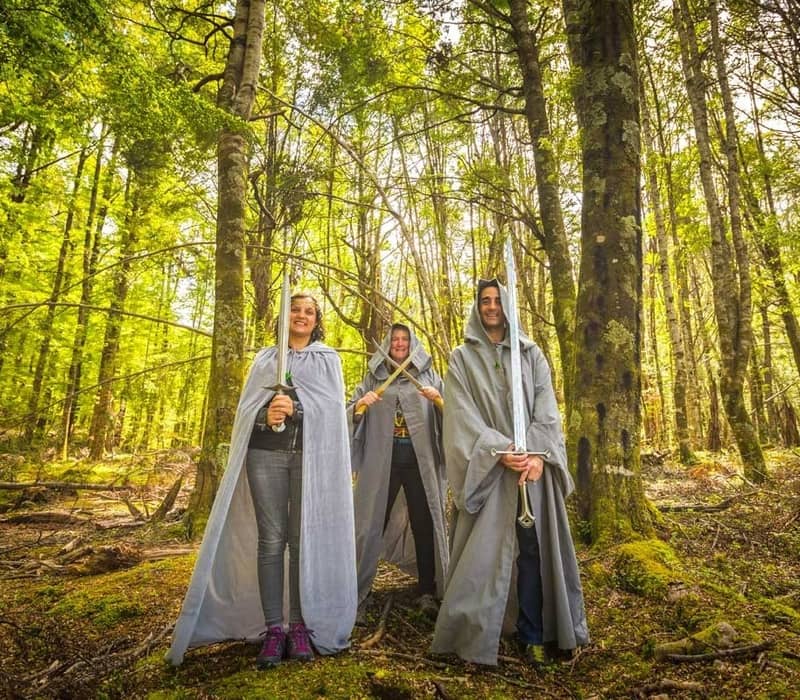 Three people wearing capes and holding swords pose in a bright green forest.