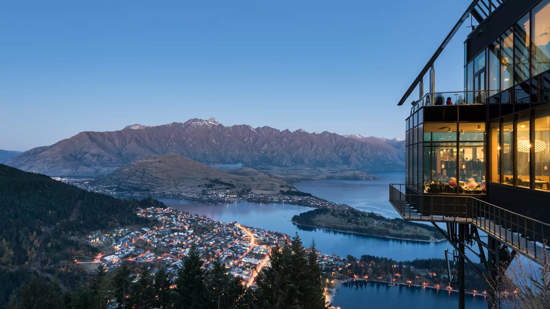View over Queenstown town, lake, and mountains with gondola chairlift in foreground under blue sky.