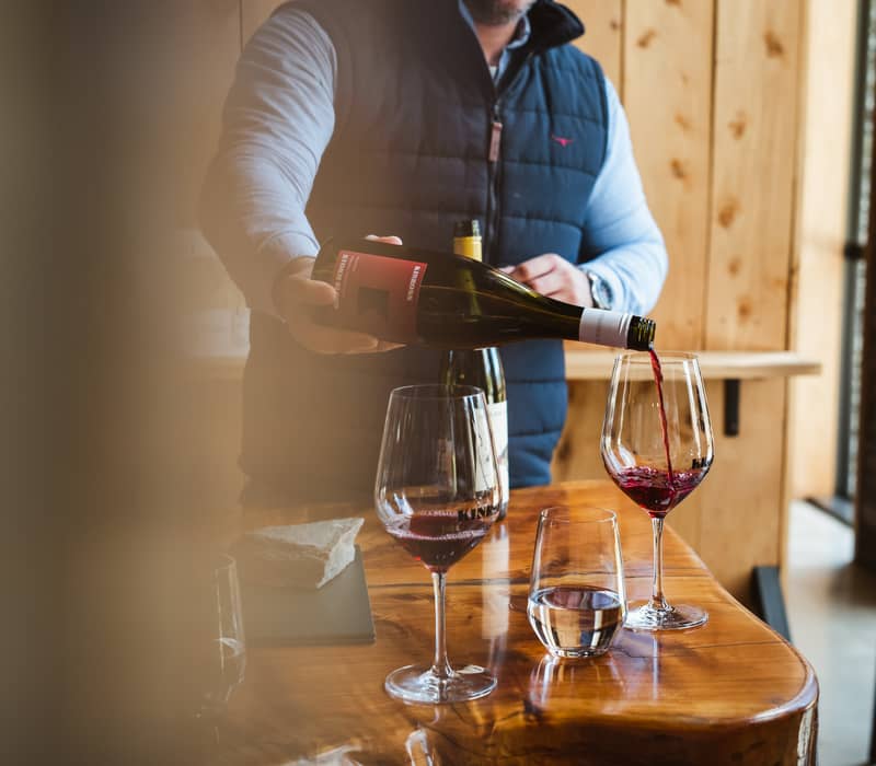 Person pouring red wine into glass on wooden table indoors.
