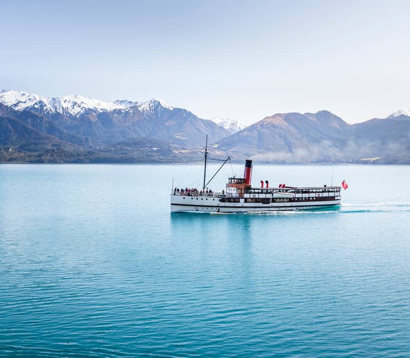 Steamboat cruising on blue lake water with snowy mountains in the background on a clear day.